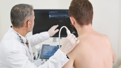 Echografist onderzoekt de schouder van een patiënt met frozen shoulder: de echokop op de schouder, het echografisch beeld zichtbaar op de monitor